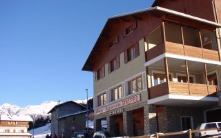 Hotel dalle linee semplice e lo stile alpino. L'inquadratura immortala l'Hotel Ginepro, con il suo tette spiovente e con dettagli come parte del rivestimento della facciate e le terrazze nel tipico legno della zona. Verso sinistra invece si intravedono i profili delle montagne innevate.