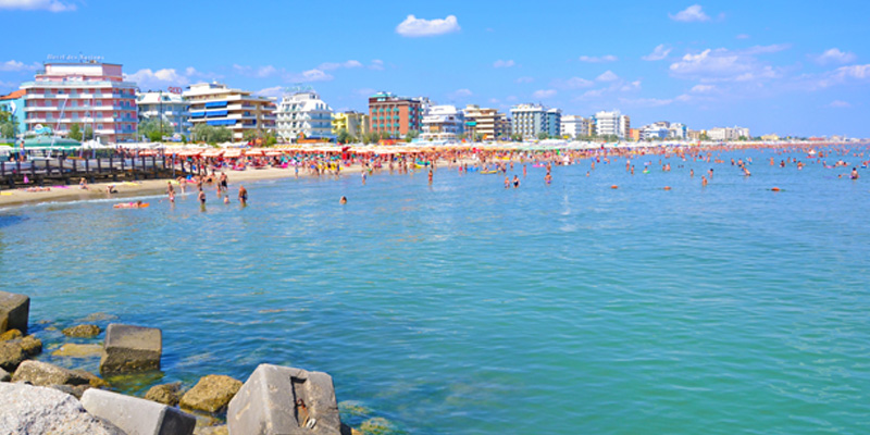 La riva di Riccione, composta da sabbia fine, abbraccia le acque blu del mare.
In lontananza si possono notare diverse strutture ricettive.