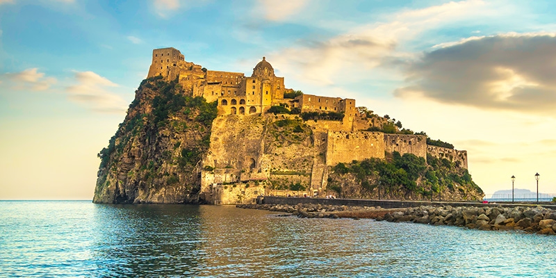 L'isola di Ischia si erge tra le acque del mare mostrando il suo antico castello, mentre viene baciata dalla luce del tramonto. La sua atmosfera romantica la rende un'ottima meta per gli innamorati.