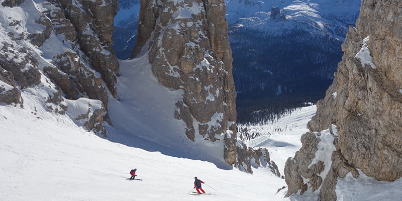 Due sciatori sciano su delle piste libere di una stretta vallata delimitata da rocce dolomitiche.