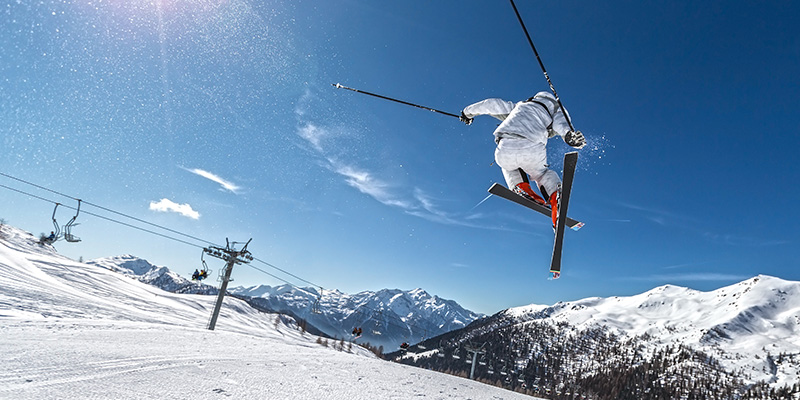 Uno sciatore è immortalato a mezz'aria in un salto sullo sci. Sullo sfondo si vedono le vette innevate delle montagne, l'impianto sciistico ed il terreno innevato della vallata.