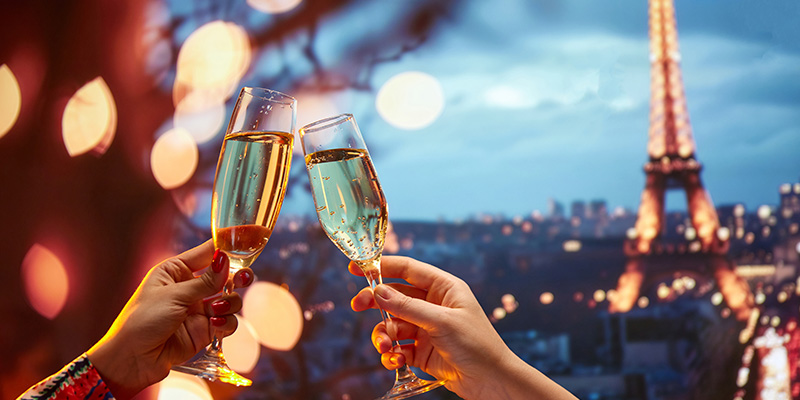 In primo piano due calici di champagne si incrociano in un brindisi Sullo sfondo si intravede la Torre Eiffel e la città, entrambe illuminate a festa!